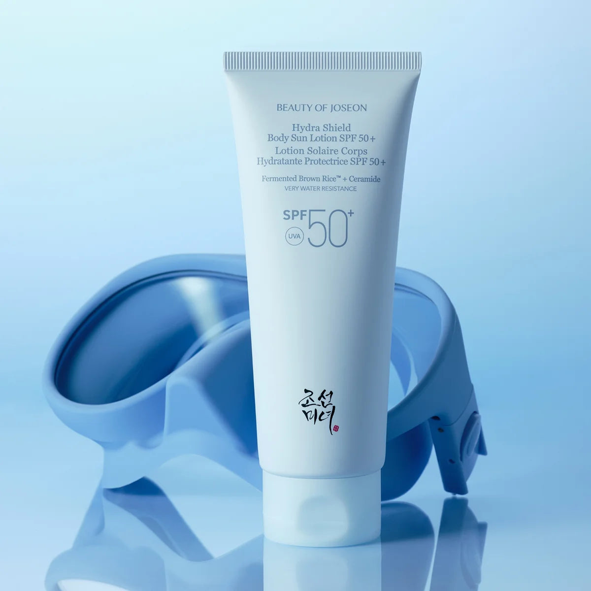 A white tube of Joseon Beauty Hydra Shield Body Sun Lotion SPF 50+ stands upright on a reflective surface, with a translucent blue swim cap in the background against a soft blue gradient backdrop.