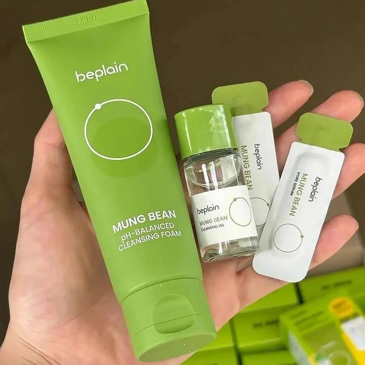 A hand holds a green Beplain Mung Bean pH-balanced cleansing foam tube alongside a small bottle of cleansing oil and two white sachets with green caps, all branded Beplain.