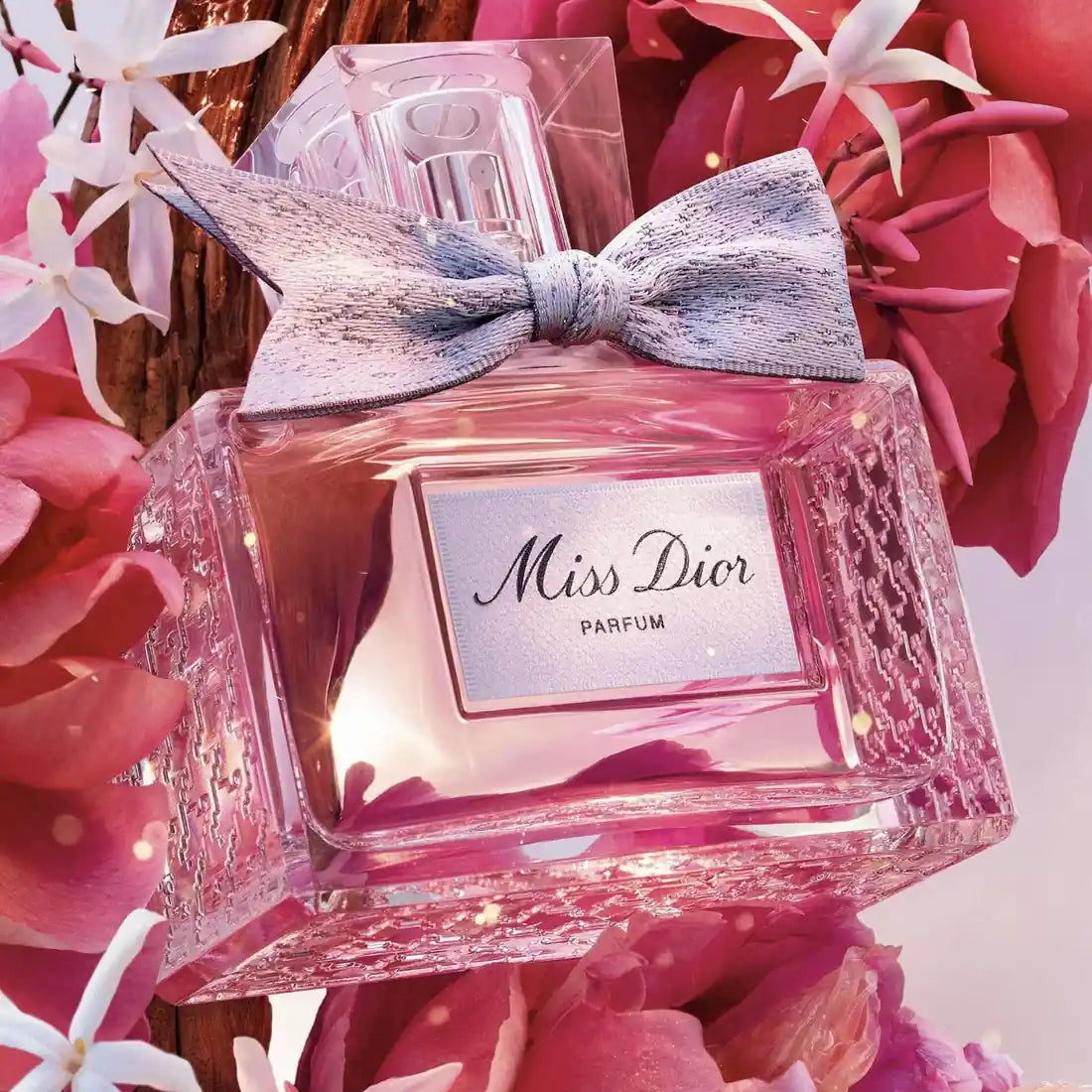 Miss Dior Parfum bottle with a pink liquid, a textured silver bow, and a silver label, nestled among pink flowers.