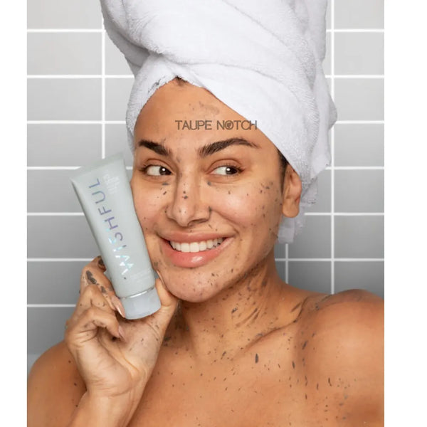 Face & body enzyme scrub tube held by person, taupe-gray; towel on head, 100 ml.