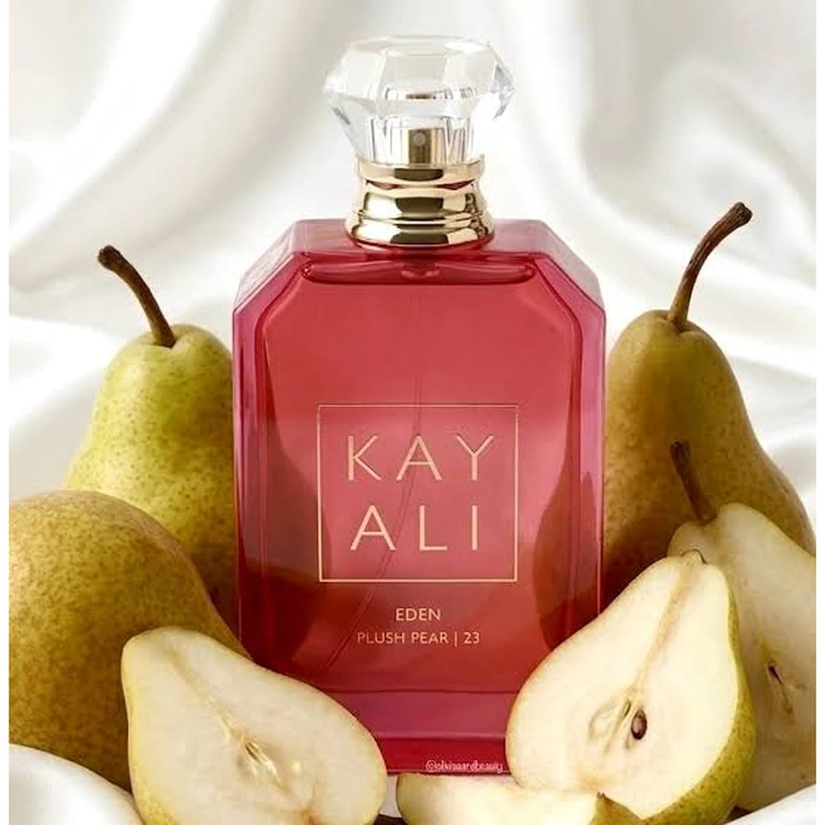 A red rectangular perfume bottle labeled KAY ALI EDEN PLUSH PEAR | 2J, surrounded by whole and sliced pears on a white draped fabric background.