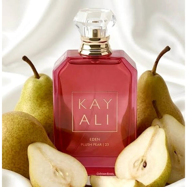 A red rectangular perfume bottle labeled KAY ALI EDEN PLUSH PEAR | 2J, surrounded by whole and sliced pears on a white draped fabric background.