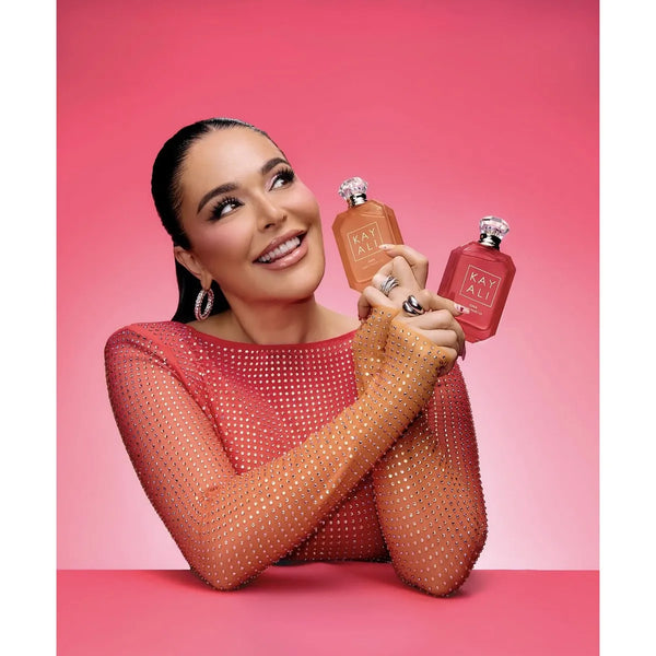 Mona kattan smiling with a sparkling peach mesh top holds two pink bottles with silver caps and a visible 'MY' logo against a pink studio background.