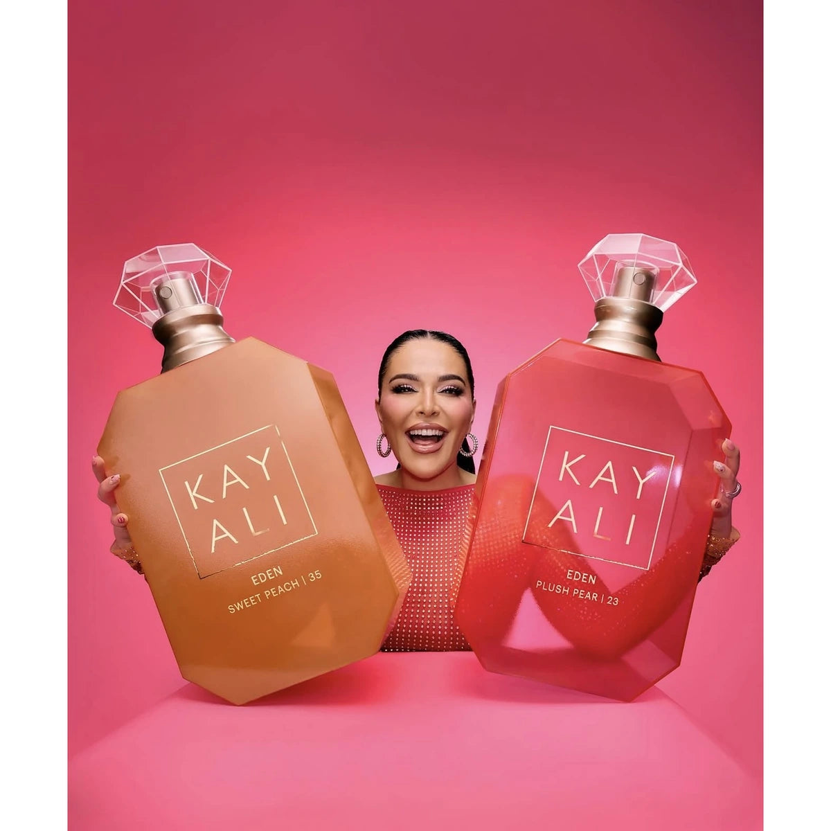 Mona kattan two large Kayali perfume bottles against a pink studio background. The bottle on the left is tan/golden and labeled Green Sweet Peach 133, and the bottle on the right is red/pink and labeled Eden Plum Pear 133.