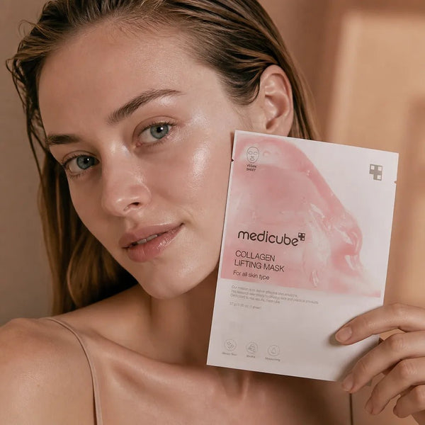 A woman holds a sheet mask package labeled 'medicube Collagen Lifting Mask' while looking at the camera. The package is white with a pink mask illustration, and the background is a softly lit warm beige indoor setting.
