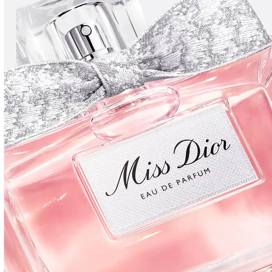 Miss Dior Eau de Parfum bottle with a pink liquid, a silver glitter bow, and a clear cap on a light gray background.