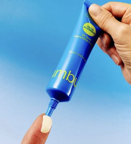A Numbuzin No.0 Zero Blemish 3X Spot Correcting Cream 20 ml 2X Spot Correcting Cream being squeezed onto a finger, with a white cream visible, against a blue gradient background.