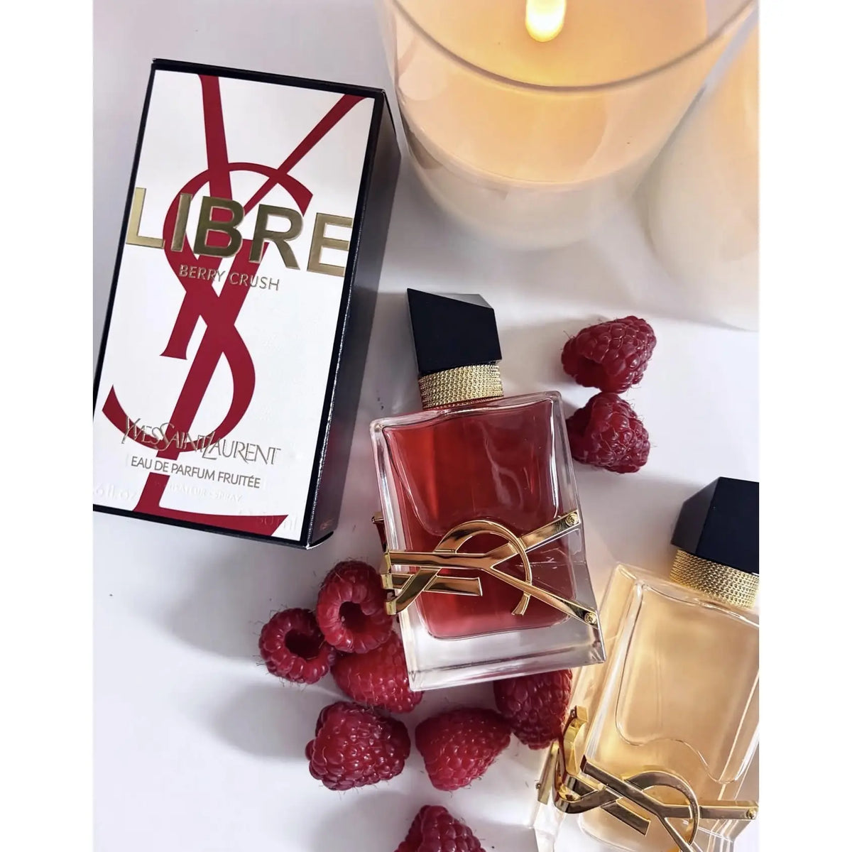 YSL Libre Berry Crush eau de parfum perfume bottles and packaging box, styled with fresh raspberries and a lit candle on a white surface.