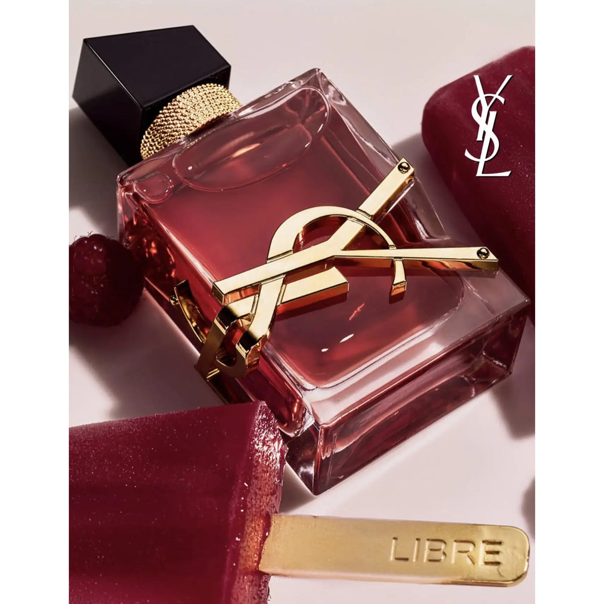 YSL Libre perfume bottle with a pinkish-red translucent glass body, gold-tone YSL logo clasp, and black cap, styled with red lip balm and a gold-toned balm stick on a light background.