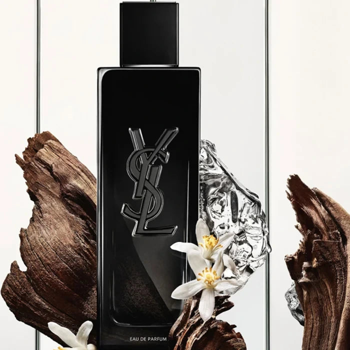 YSL Eau de Parfum bottle in glossy black glass with a silver interlocking YSL logo, styled with white flowers, textured wood, and a clear crystal fragment against a light neutral background.