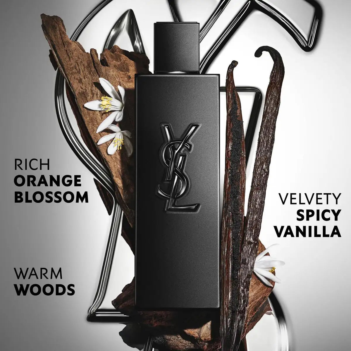 YSL Rich Orange Blossom Eau de Parfum bottle in matte black, styled with vanilla pods, white flowers, and wood pieces inside a wire frame. Text on the left reads 'RICH ORANGE BLOSSOM' and text on the right reads 'VELVETTY SPICY VANILLA' with 'WARM WOODS' below.